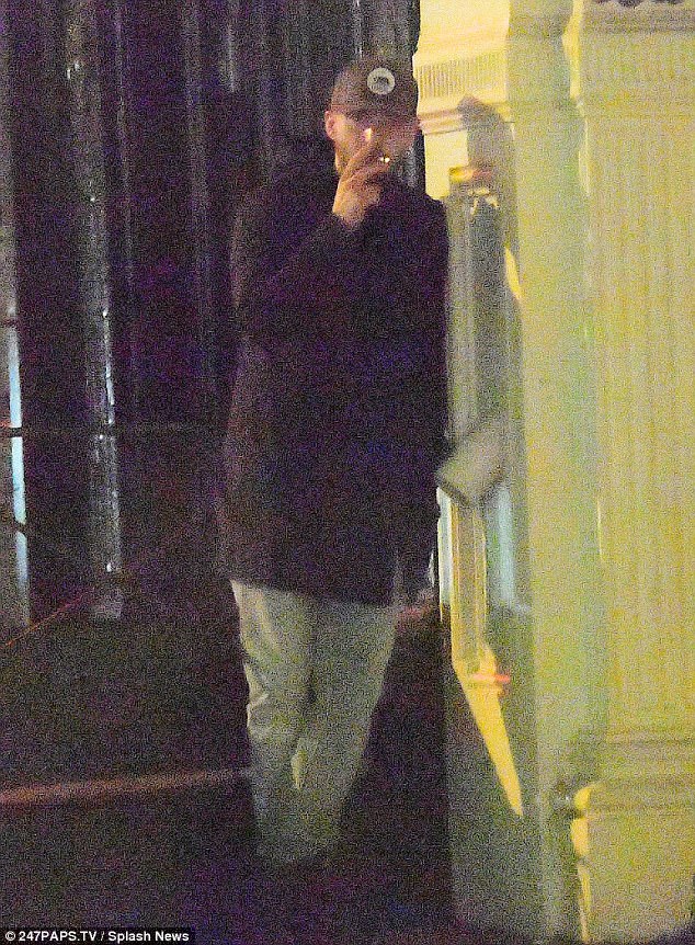 Intermedio: Justin fue apurado apoyado contra el edificio fumando un cigarrillo.  Llevaba un abrigo oscuro sobre pantalones holgados y una gorra de béisbol.